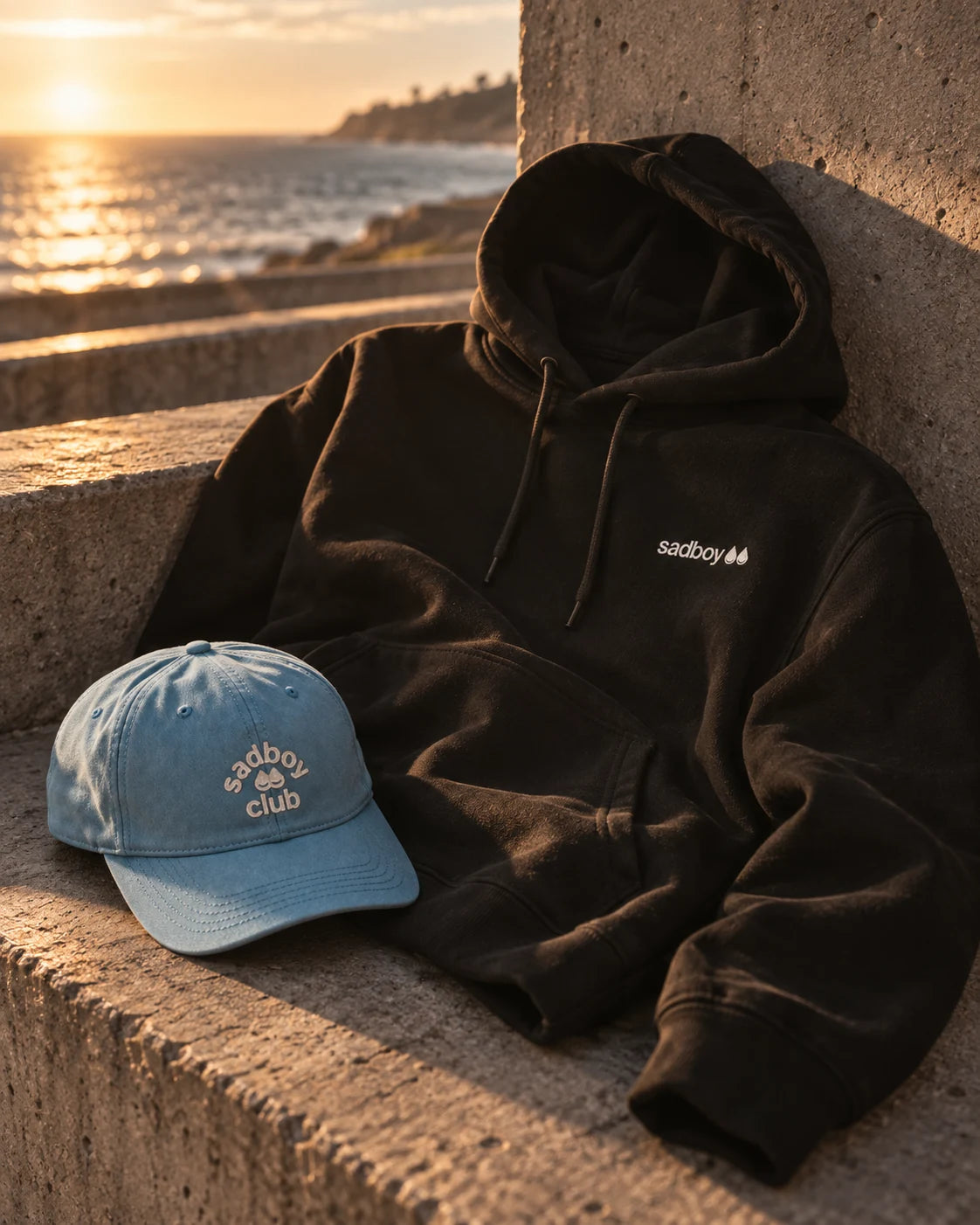 Black Sad Boy hoodie and powder-blue cap resting on concrete
